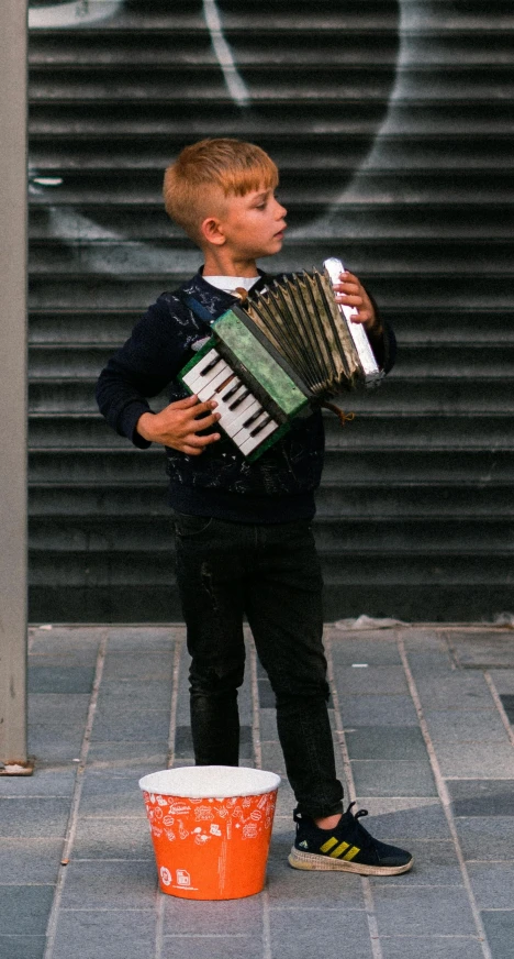 a young boy is playing an accordion on the sidewalk, by Lucia Peka, pexels contest winner, square, 15081959 21121991 01012000 4k, standing upright, chilean