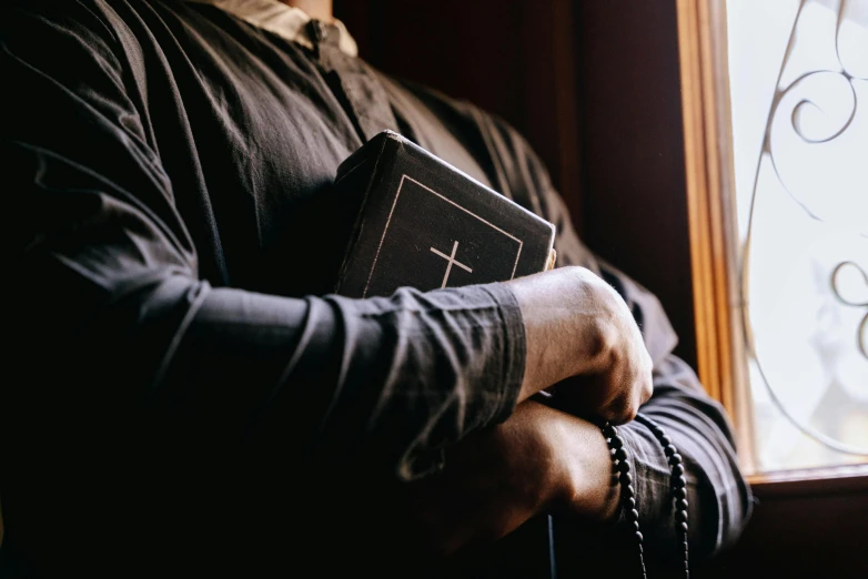 a man holding a bible in front of a window, unsplash, wearing cross on robe, black, instagram post, uploaded