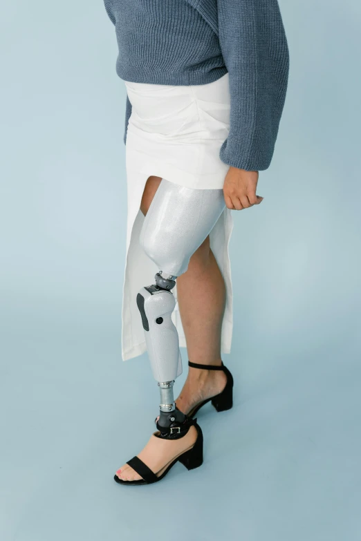 a woman with a prosthetic leg standing in front of a blue background, grey metal body, an all white human, knee, casually dressed