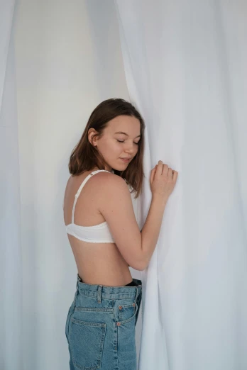 a woman standing in front of a white curtain, wearing bra, sydney sweeney, relaxed posture, facing away from camera