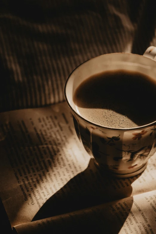 a cup of coffee sitting on top of an open book, a black and white photo, unsplash, romanticism, bathed in golden light, 2000s photo, newspaper, sepia colors