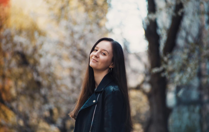 a woman in a black jacket standing in front of a tree, pexels contest winner, girl with dark brown hair, profile image, smiling, avatar image