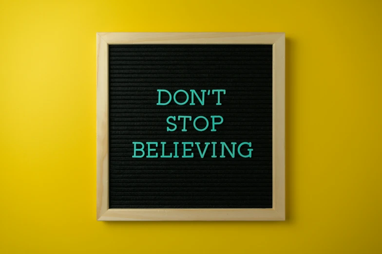 a sign that says don't stop believing, a poster, shutterstock, taken on iphone 1 3 pro, black. yellow, ethics, black and teal paper