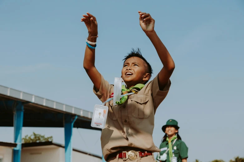 a boy in a scout uniform is flying a kite, a portrait, pexels contest winner, happening, holding a bottle of arak, cheering, thawan duchanee, brown