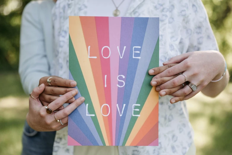 a person holding a sign that says love is love, poster art, by Julia Pishtar, pexels, private press, multi - coloured, greeting card, magazine, religious