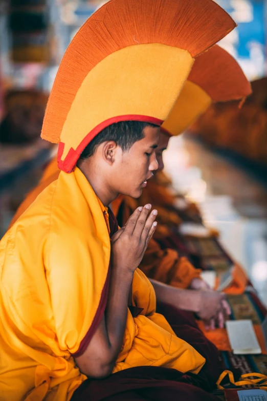 a person that is sitting down with a hat on, monk's robe, yellow and orange color scheme, praying, square