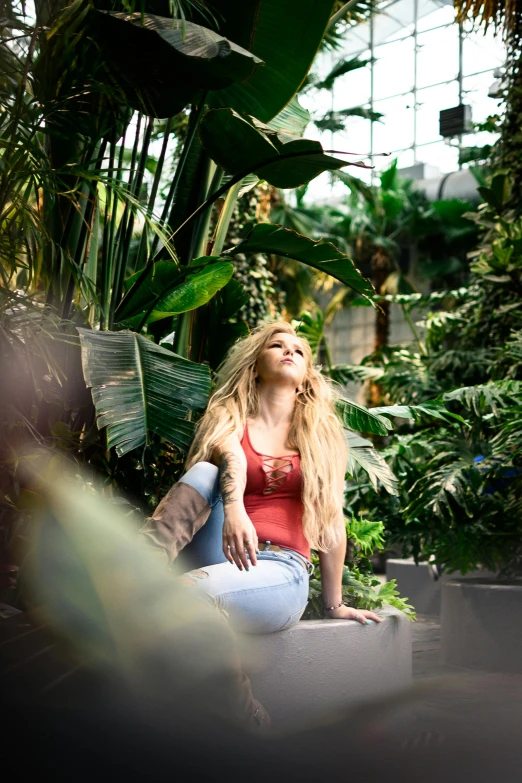 a woman sitting on a ledge in a garden, inspired by Elsa Bleda, pexels contest winner, long hair and red shirt, biodome, tropical plants, very very long blond curly hair