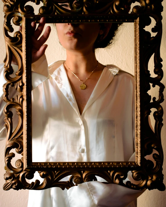 a woman holding a picture frame in front of her face, an album cover, inspired by Elsa Bleda, trending on pexels, renaissance, gold necklace, wearing a white button up shirt, on a reflective gold plate, francesca woodman style