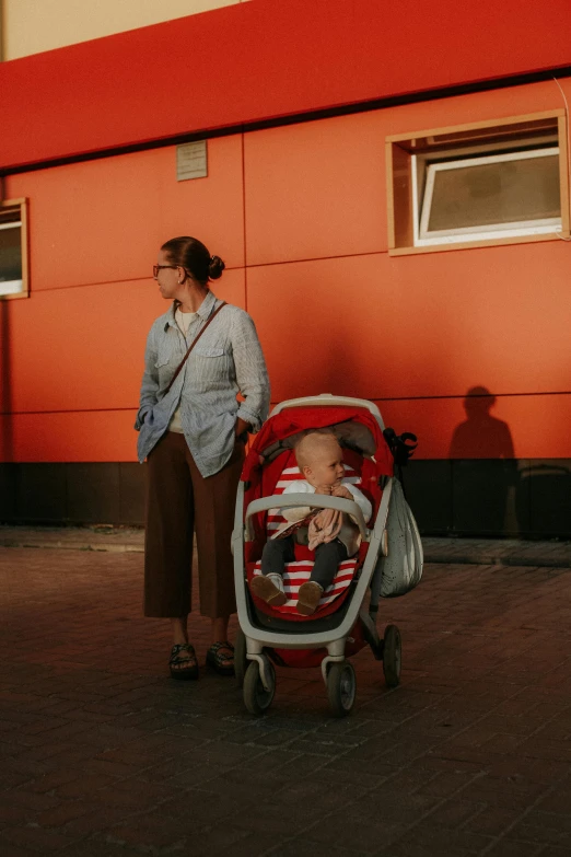 a woman standing next to a baby in a stroller, by Attila Meszlenyi, pexels contest winner, symbolism, red and brown color scheme, summer evening, square, serious business