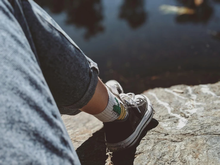 a person sitting on a rock next to a body of water, trending on pexels, photorealism, striped socks, with a tree in the background, wearing white sneakers, green legs