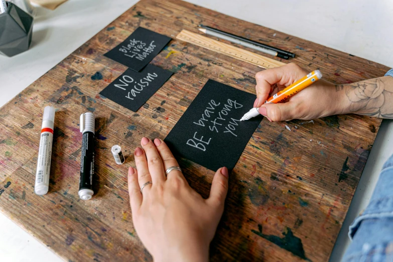 a person writing on a piece of wood, by Julia Pishtar, trending on unsplash, arbeitsrat für kunst, matte black paper, placards, molotow premium color palette, pair of keycards on table