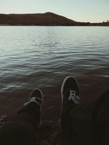 a person sitting on a beach next to a body of water, inspired by Elsa Bleda, sneaker photo, trending on vsco, it has a lake in the distance, it's getting dark