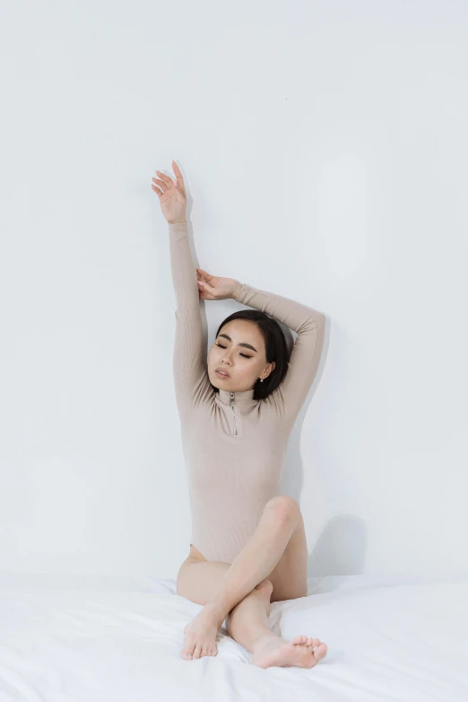 a woman sitting on top of a bed next to a white wall, inspired by Elizabeth Polunin, trending on unsplash, arabesque, wearing turtleneck, muted colored bodysuit, with arms up, charli xcx