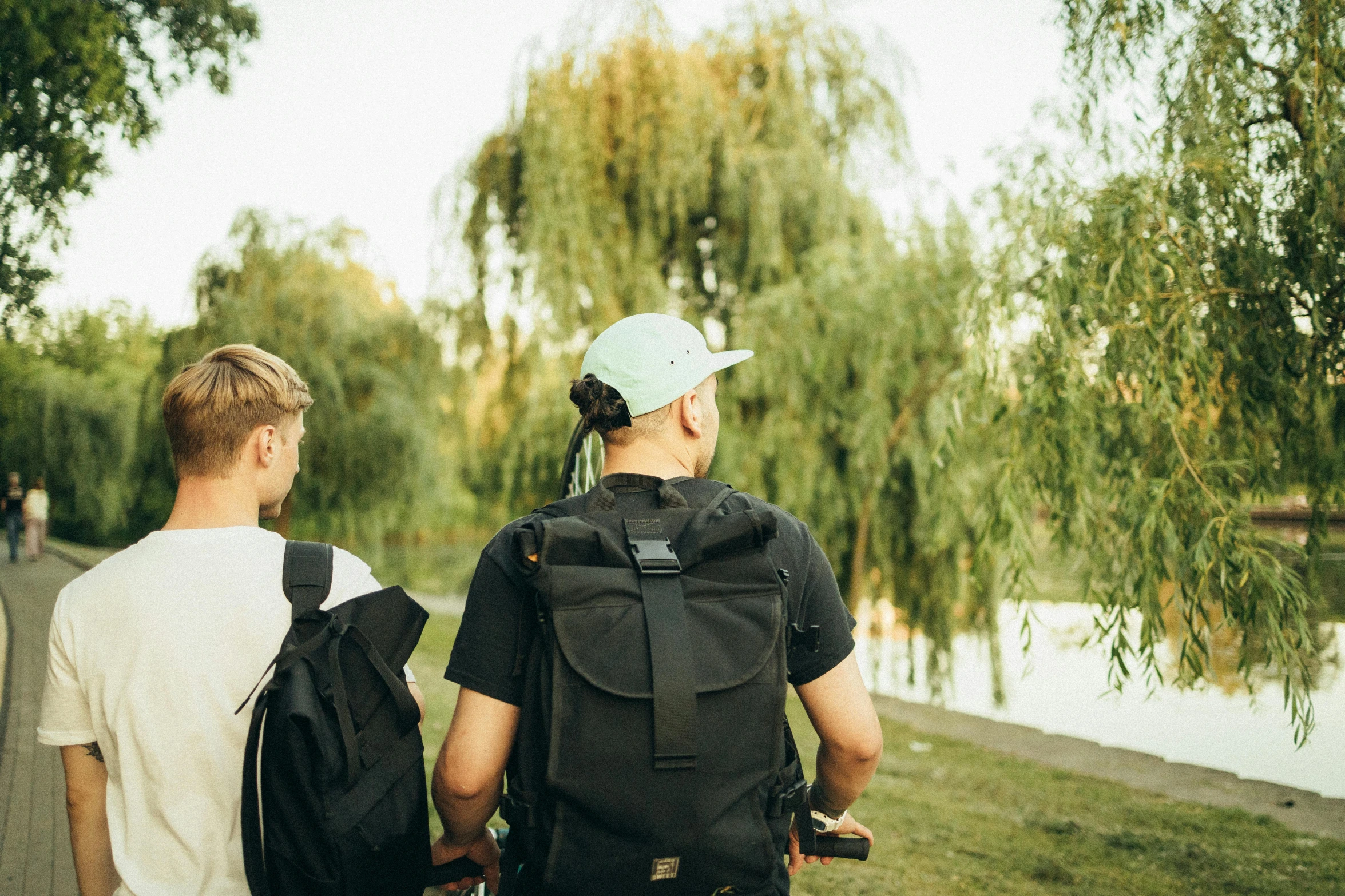 two men walking down a path next to a body of water, pexels, a backpack, willow trees, avatar image, sports photo