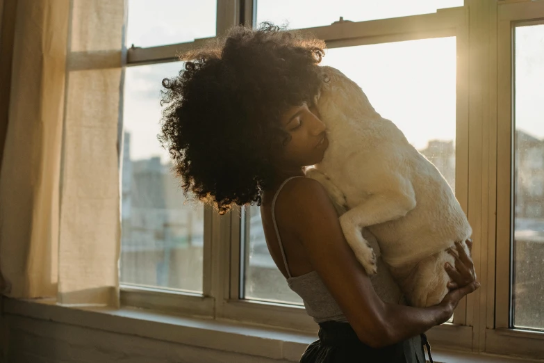 a woman holding a cat in front of a window, pexels contest winner, subject: dog, african american woman, beautiful late afternoon, holding each other