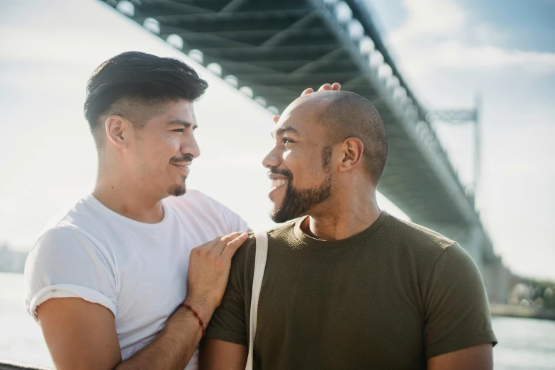 two men standing next to each other near a bridge, mix of ethnicities and genders, muscular men entwined together, profile image, smiling at each other