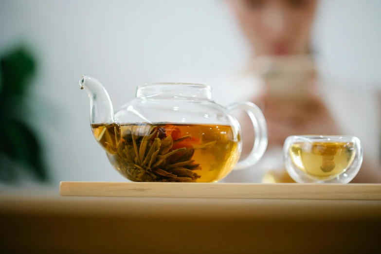 a tea pot sitting on top of a table next to a cup of tea, trending on pexels, with clear glass, person in foreground, botanicals, thumbnail