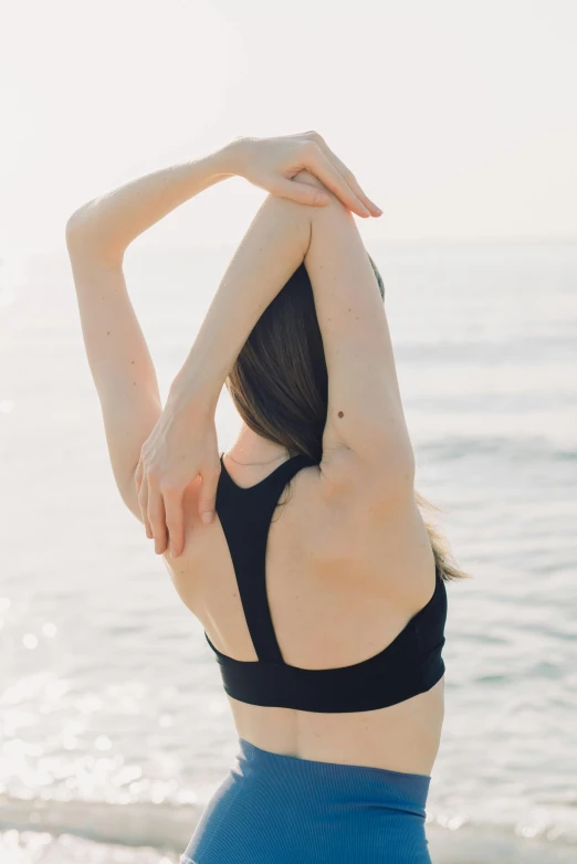 a woman standing on top of a beach next to the ocean, a tattoo, unsplash, renaissance, sports bra, raised hand, bent over posture, black