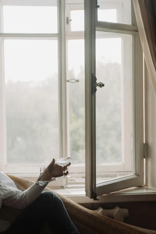 a woman sitting on a couch looking out a window, holding a drink, overcast day, ignant, photo of a beautiful window