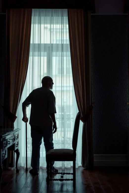 a man standing in front of a window next to a chair, walking away from the camera, nursing home, hotel room, movie still