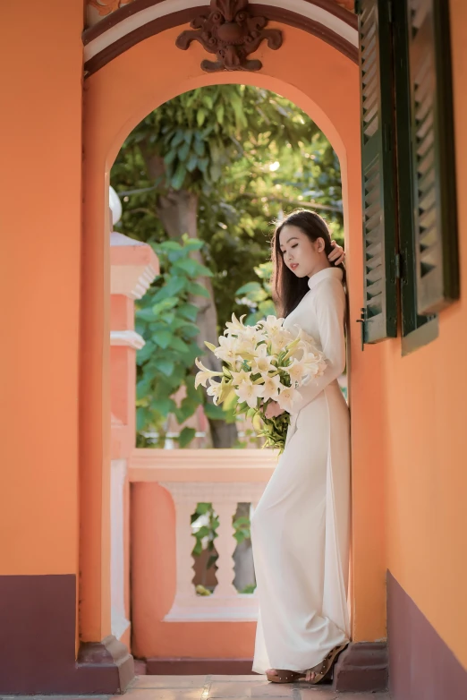 a woman in a white dress holding a bouquet, a picture, inspired by Ruth Jên, archways, orange and white color scheme, ao dai, outside view