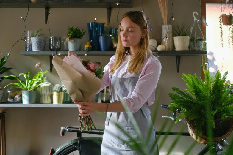 a woman standing next to a bicycle holding a bunch of flowers, a still life, by Alice Mason, pexels contest winner, wearing an apron, flower shop scene, still from the film, mid morning lighting