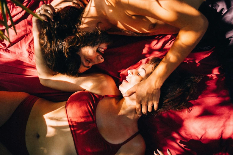 a woman laying on top of a bed next to a man, trending on pexels, romanticism, wearing red clothes, woman holding another woman, in a sunbeam, two muscular men entwined