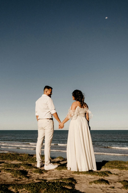 a man and a woman holding hands on a beach, a picture, pexels contest winner, in a long white dress, looking towards the horizon, blue skies, overlooking