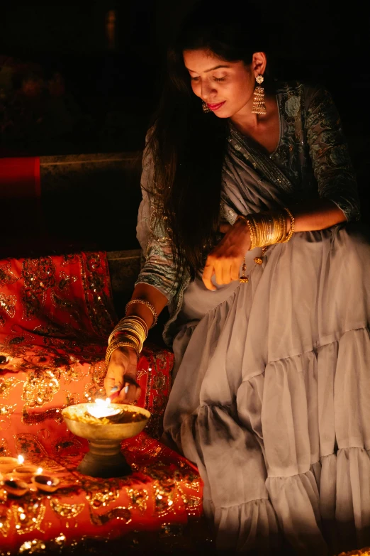 a woman sitting on the ground lighting a candle, trending on pexels, renaissance, wearing a silk kurta, in a glowing skirt, festive atmosphere, grey