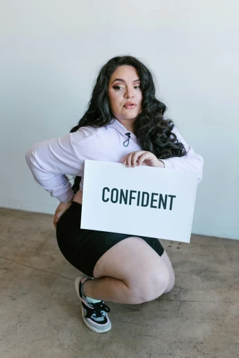 a woman kneeling down holding a sign that says confident, an album cover, bbw, self-confidence, kailee mandel, wearing tight simple clothes