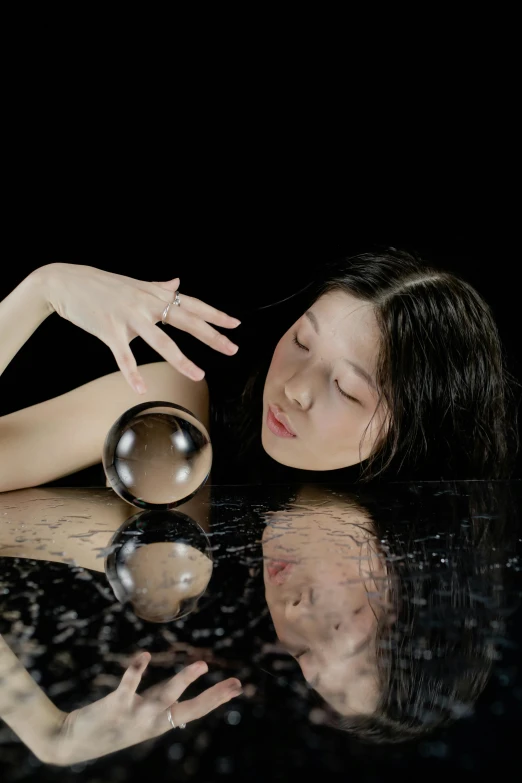 a woman laying on top of a table next to a glass ball, an album cover, inspired by Kanō Naizen, unsplash, partially cupping her hands, mirror like water, female magician, studio photo