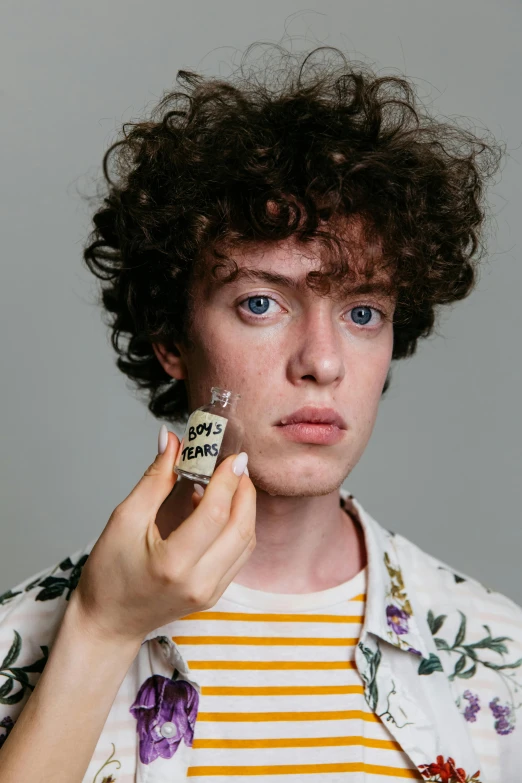 a close up of a person holding a bottle of perfume, an album cover, inspired by Grayson Perry, trending on pexels, 1 7 - year - old boy thin face, curly haired, sophia lillis, studio portrait