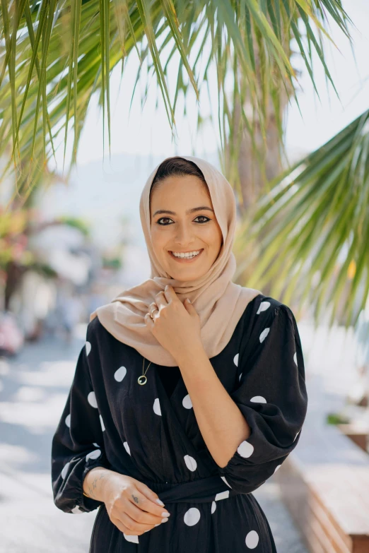 a woman wearing a black and white polka dot dress, inspired by Maryam Hashemi, pexels contest winner, hurufiyya, smiling at camera, with palm trees in the back, wearing a head scarf, wearing casual clothing