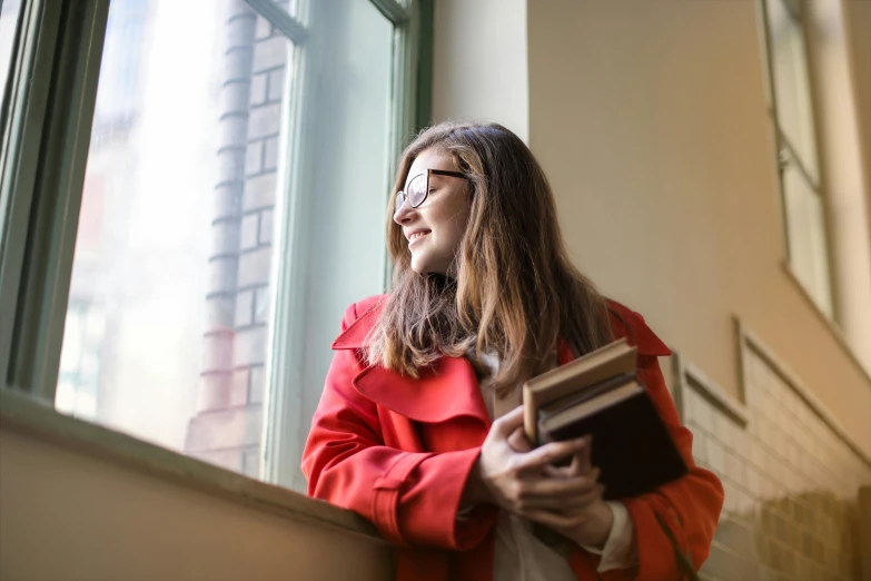 a woman standing by a window holding a book, happening, wearing red jacket, post graduate, textbooks and books, girl with brown hair