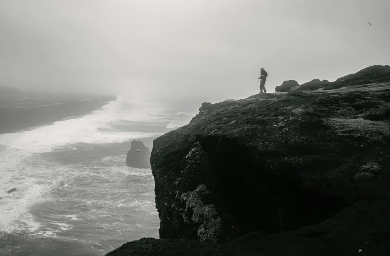 a man standing on top of a cliff next to the ocean, a black and white photo, by Kristian Zahrtmann, misty weather, low quality photo, trending photo, landscape photo