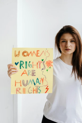 a woman standing in front of a white wall holding a sign, a poster, by Julia Pishtar, trending on pexels, feminist art, her skin is light brown, hr, a colorful, background image