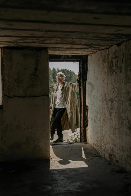 a man standing in a doorway of a building, an album cover, unsplash, white haired, wearing trenchcoat, in an abandoned barn, style of ilya kushinov