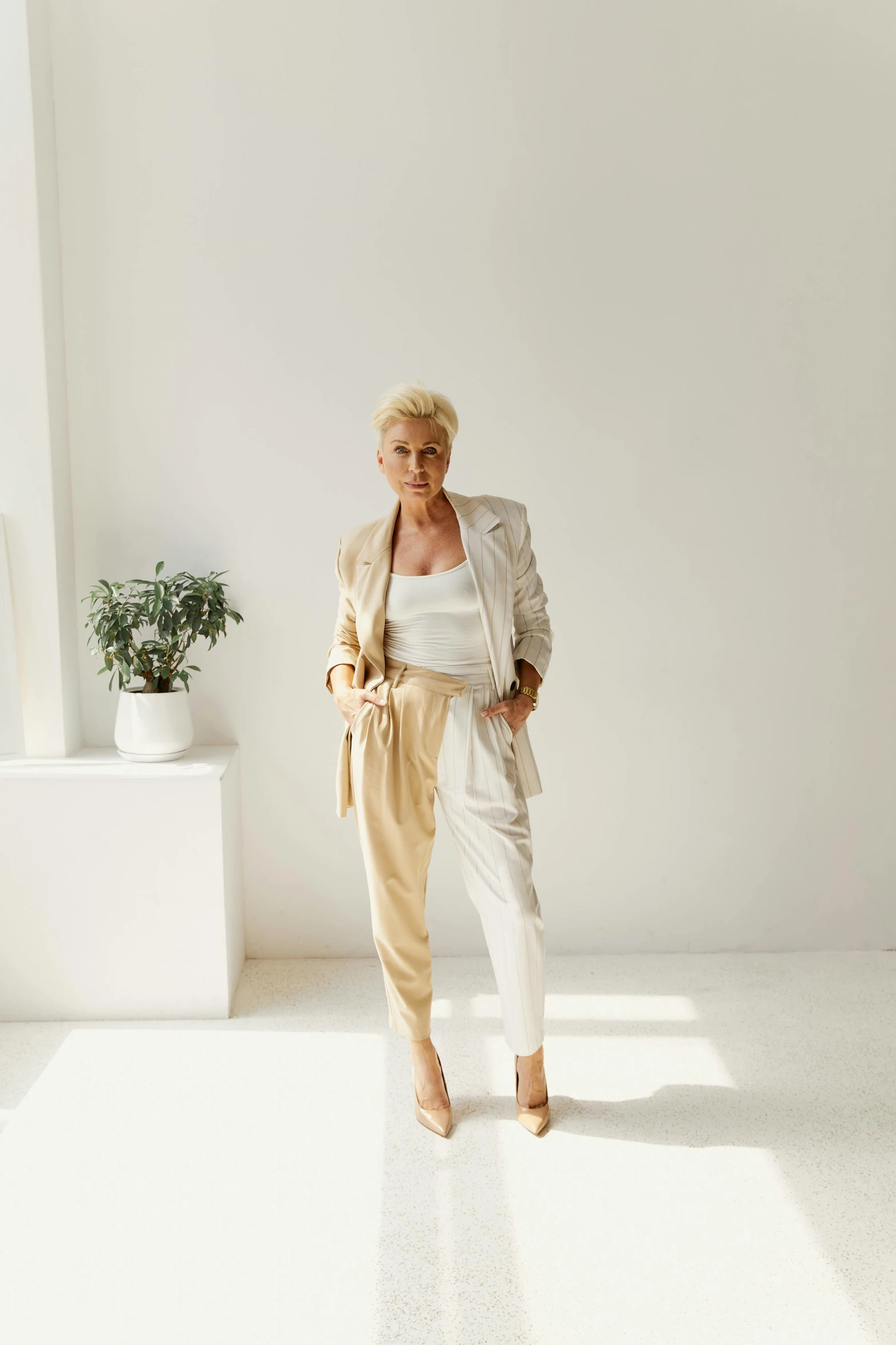 a woman standing in a white room next to a window, by Ellen Gallagher, trending on unsplash, gold suit jacket in snake print, bleached blonde short hair, white pants, three piece suit