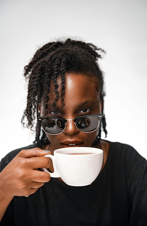 a woman sitting at a table with a cup of coffee, by Lily Delissa Joseph, trending on unsplash, afrofuturism, in front of white back drop, wearing shades, attractive man drinking coffee, androgynous face