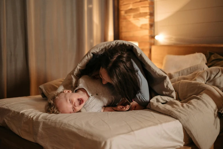 a woman laying on top of a bed next to a baby, pexels contest winner, happening, curled up under the covers, australian, head bent back in laughter, evening lighting