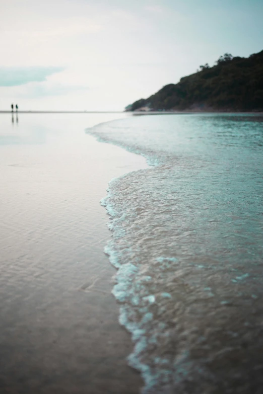 a couple of people standing on top of a sandy beach, pristine rippling oceanic waves, muted colours, up-close, beaches