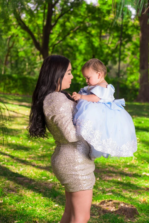 a woman holding a baby in her arms, instagram, wearing silver dress, latinas, slide show, lush surroundings