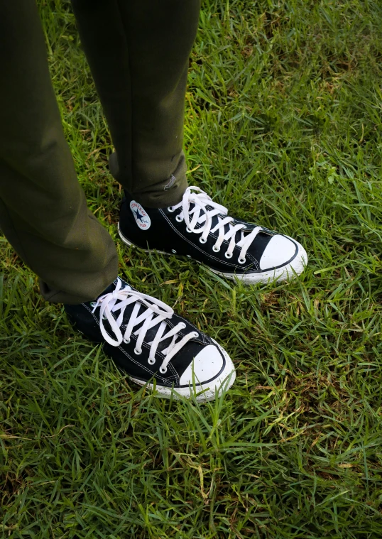 a person standing on top of a lush green field, wearing white sneakers, black sweatpants, converse, zoomed in
