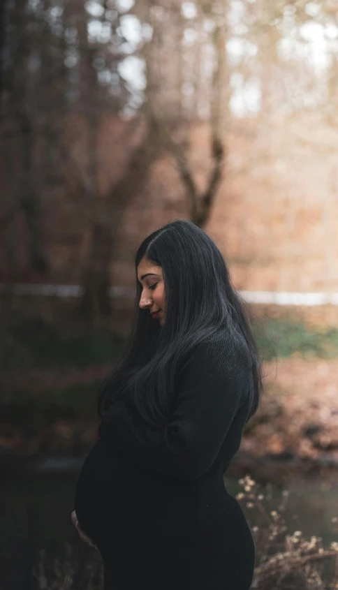 a pregnant woman standing in front of a river, an album cover, inspired by Elsa Bleda, pexels contest winner, realism, long free black straight hair, wearing a black sweater, thoughtful expression, in a city park