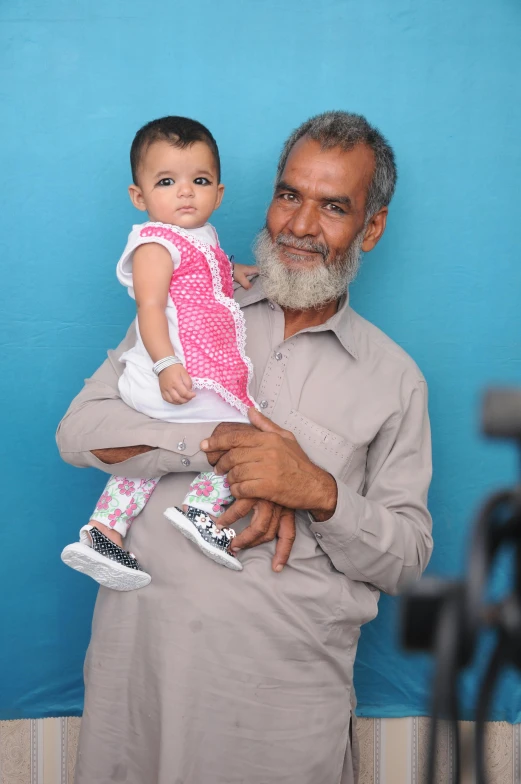 a man holding a baby in his arms, by Riza Abbasi, pexels contest winner, cyan photographic backdrop, grandfatherly, looking to camera, in 2 0 1 5