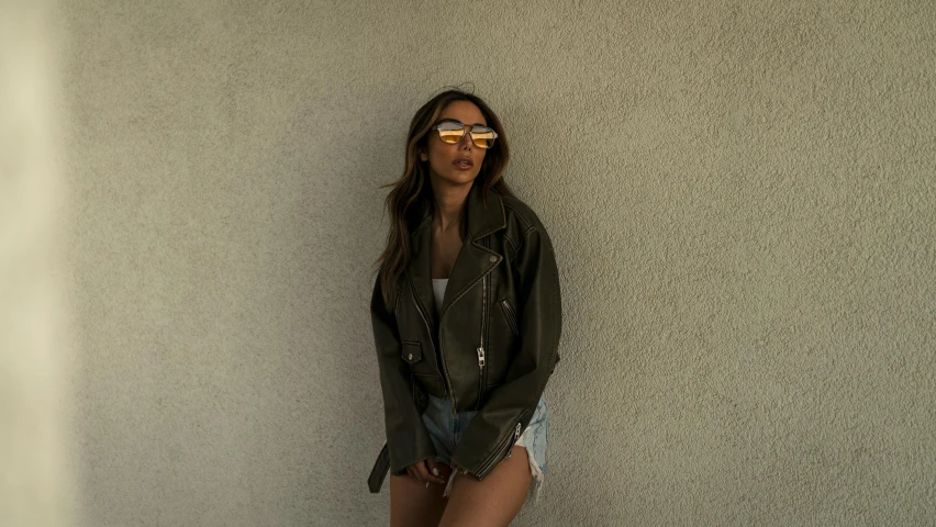 a woman leaning against a wall wearing sunglasses, by Robbie Trevino, madison beer, low quality photograph, in an old 1950s leather jacket, an olive skinned