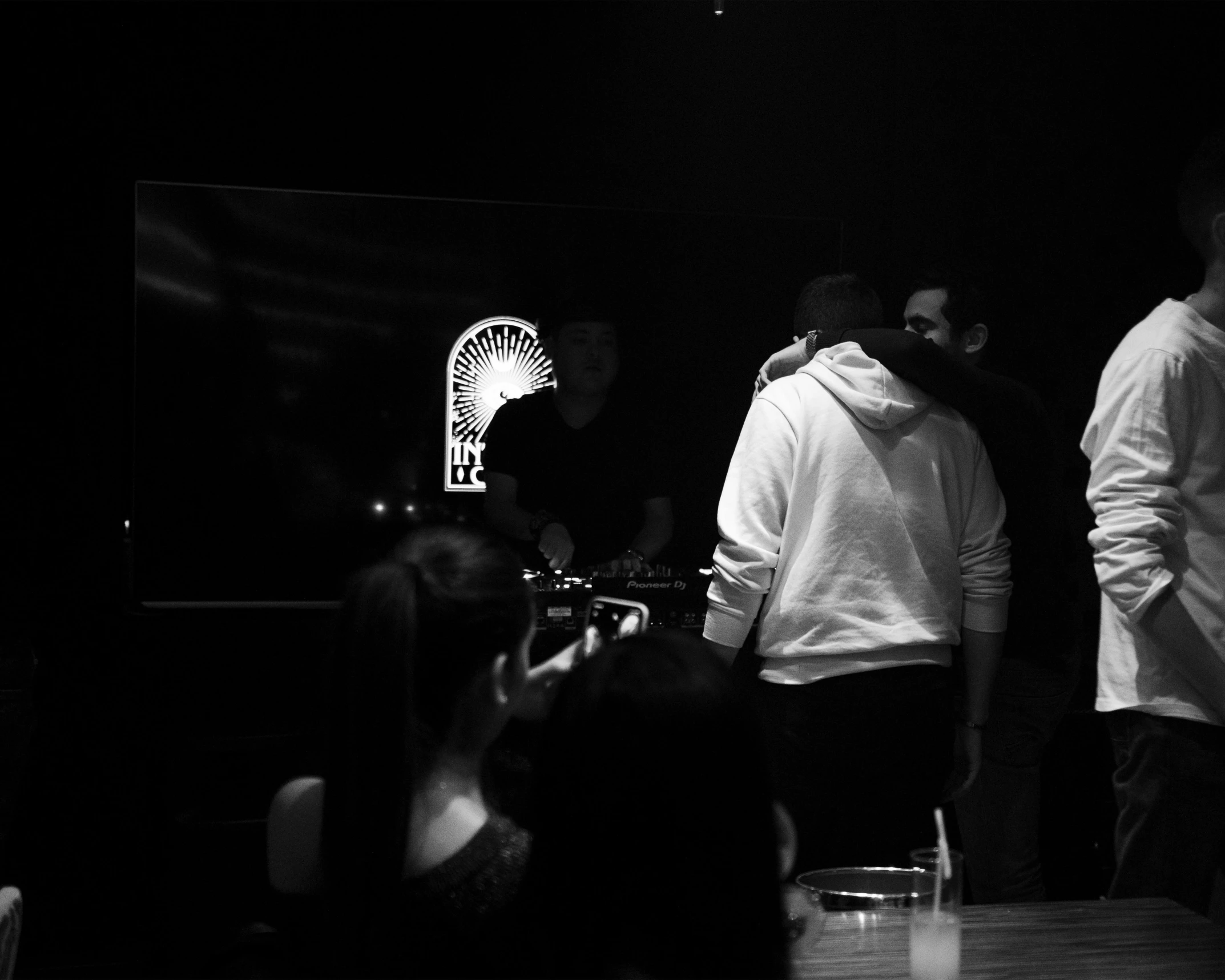 a group of people standing around a wooden table, a black and white photo, unsplash, hurufiyya, reflecting light in a nightclub, secret cypher, vantablack wall, instagram story