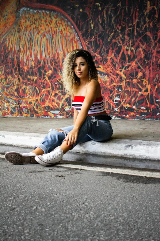 a woman sitting on a curb in front of a graffiti wall, an album cover, inspired by Esaias Boursse, trending on pexels, croptop, curly haired, casual pose, color block