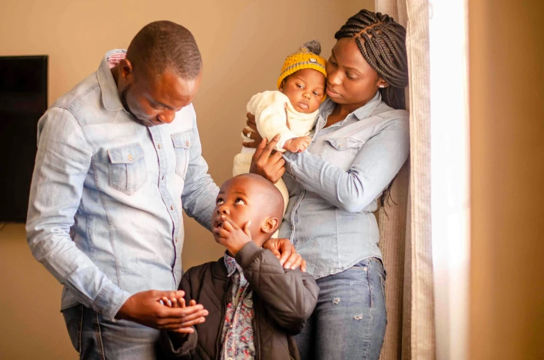 a man standing next to a woman holding a baby, by Ingrida Kadaka, pexels, visual art, sickness, husband wife and son, touching heads, mongezi ncaphayi