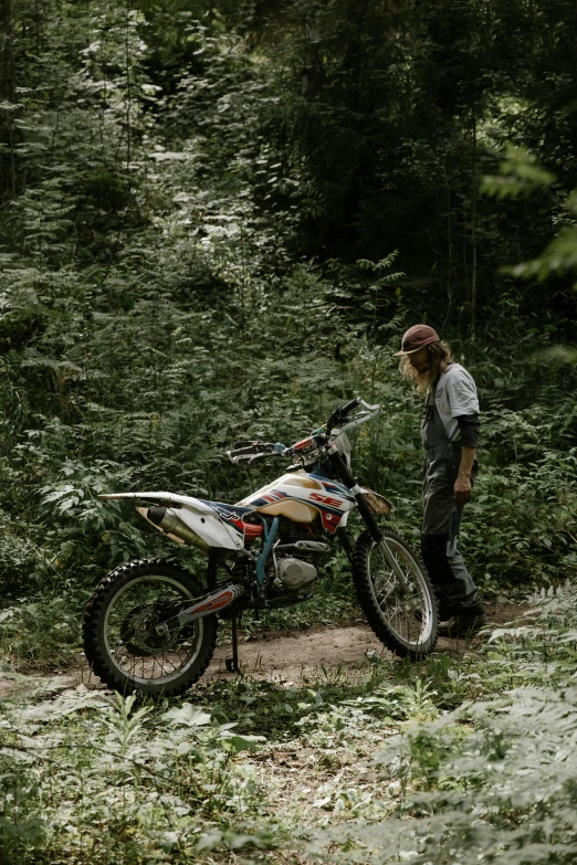 a man standing next to a dirt bike in the woods, a picture, unsplash, renaissance, 2 5 6 x 2 5 6 pixels, looking off to the side, terrain, next to a plant